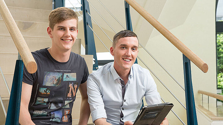 zwei Studenten sitzten auf einer Treppe, schauen in die Kamera und halten ein Tablet in der Hand