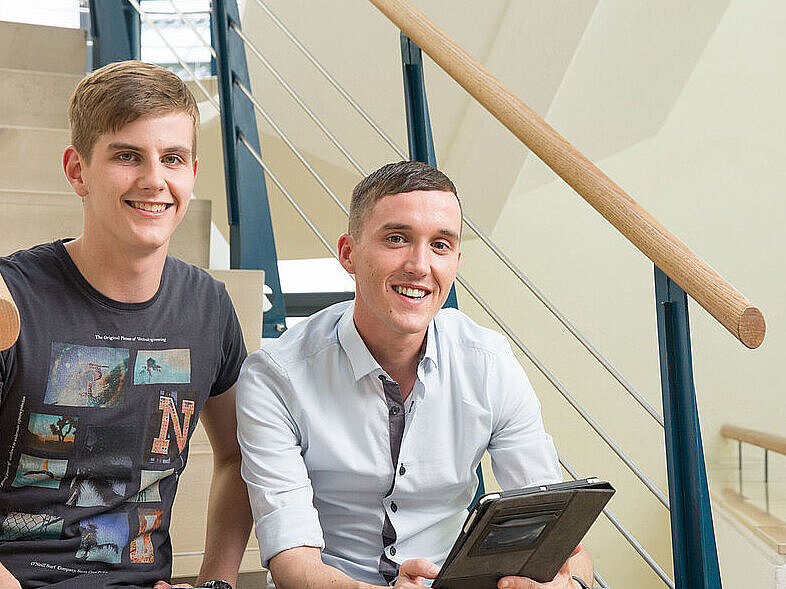 zwei junge Männer sitzen auf einer Treppe, lächeln in die Kamera und haben ein Tablet in der Hande