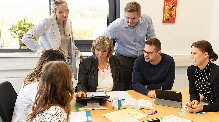 fünf Frauen und Männer diskutieren sitzend um einen Schreibtisch mit Büchern, Laptop und Schreibutensilien