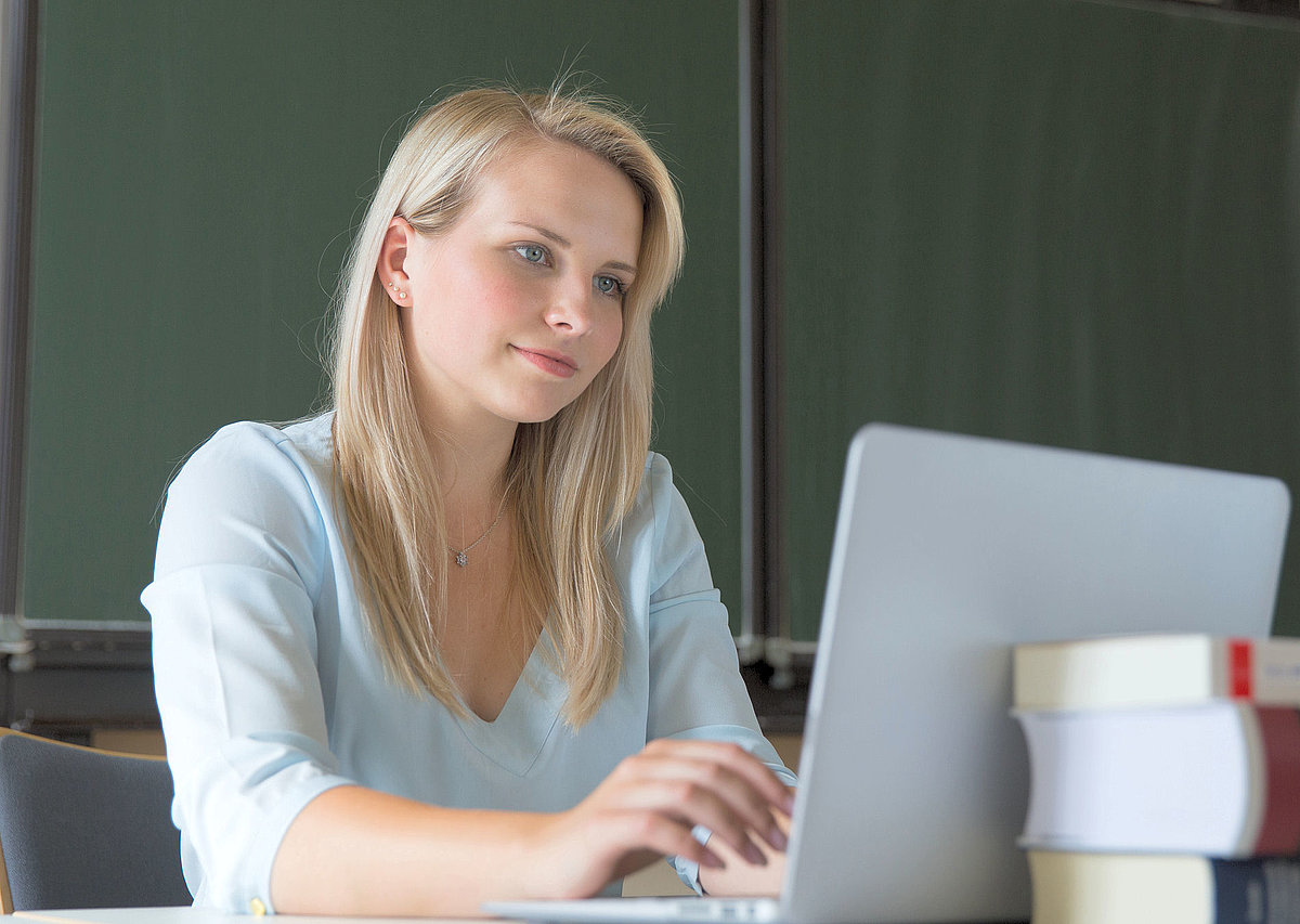 Junge blonde Frau sitzt am Schreibtisch und schaut in den Laptop.