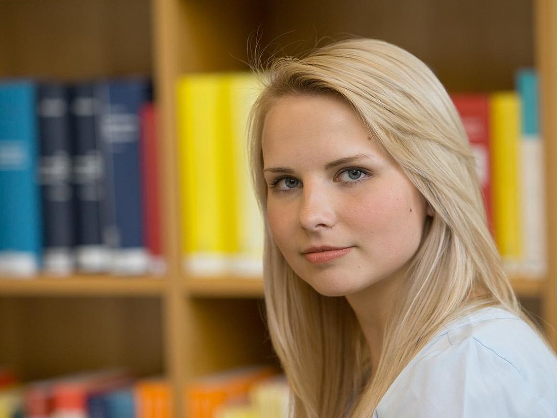 Studentin schaut in die Kamera. Im Hintergrund steht ein Bücherregal.