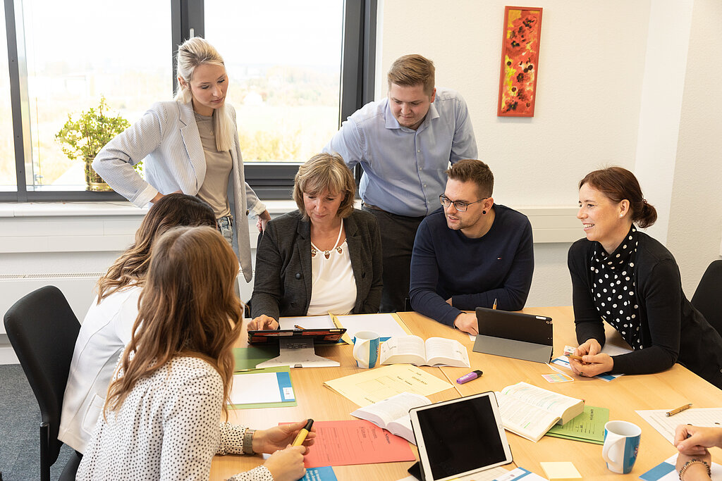fünf Frauen und Männer diskutieren sitzend um einen Schreibtisch mit Büchern, Laptop und Schreibutensilien