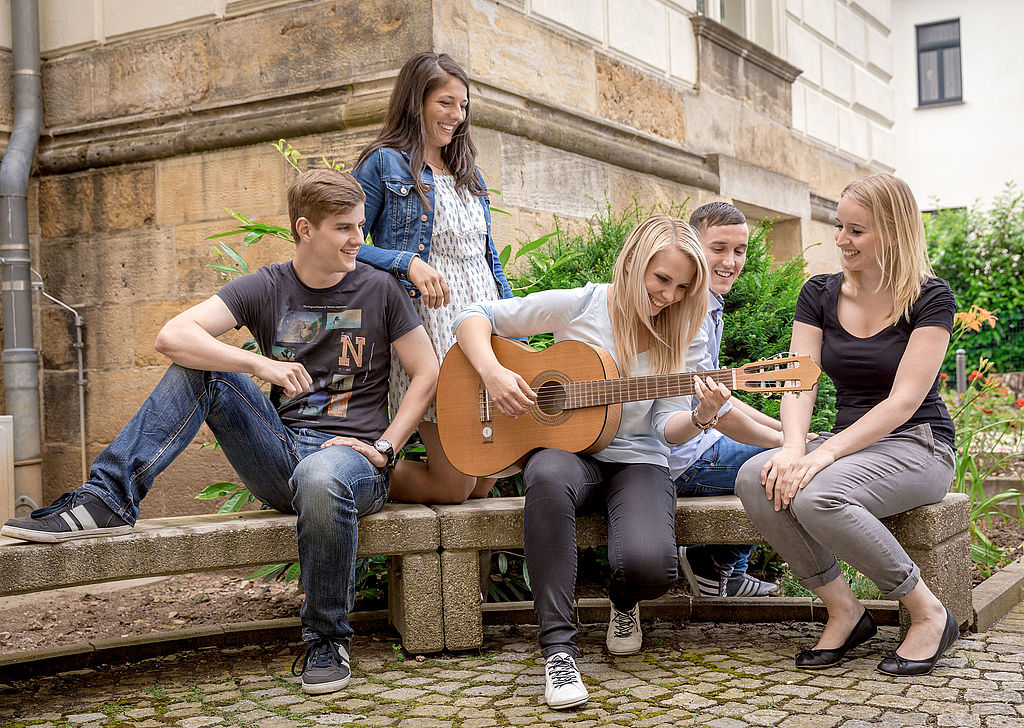 eine Gruppe von Studenten sitzen auf einer Bank und lauschen einer Musikerin mit Gitarre